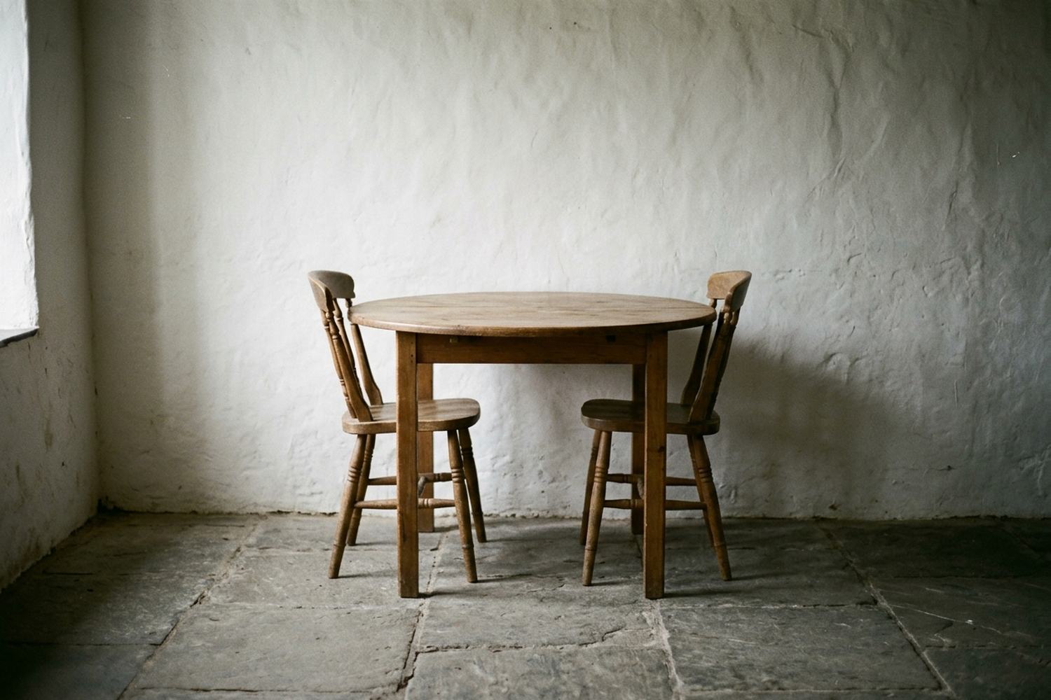 A plain wooden table with two chairs pushed in, against a bare wall.