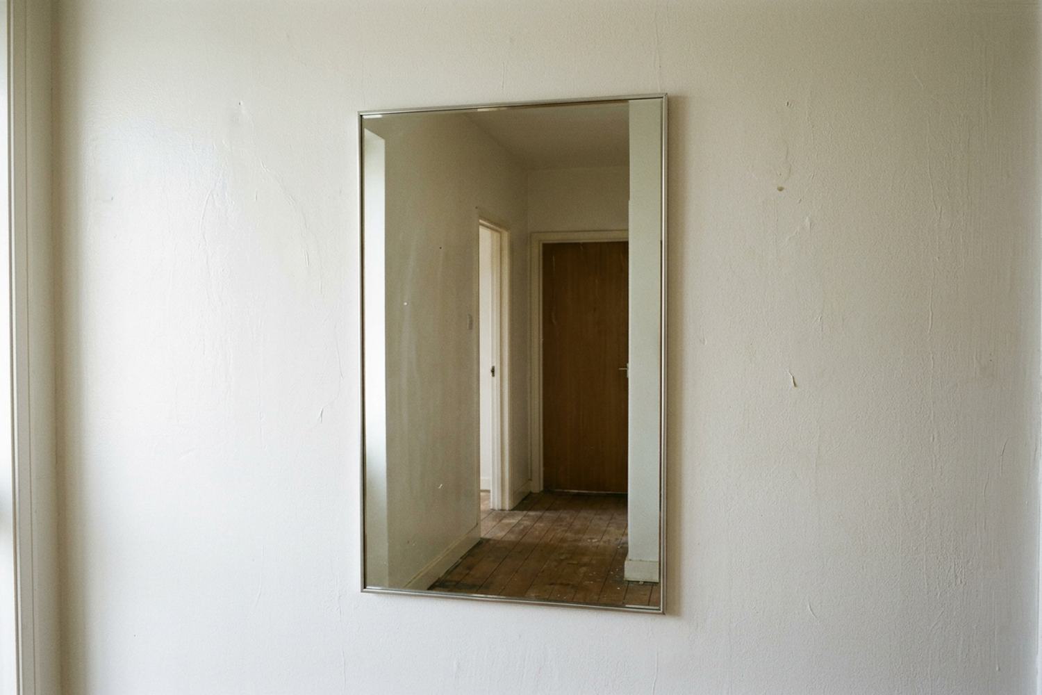 A hall mirror on a bare wall reflecting an empty hallway and a doorway.